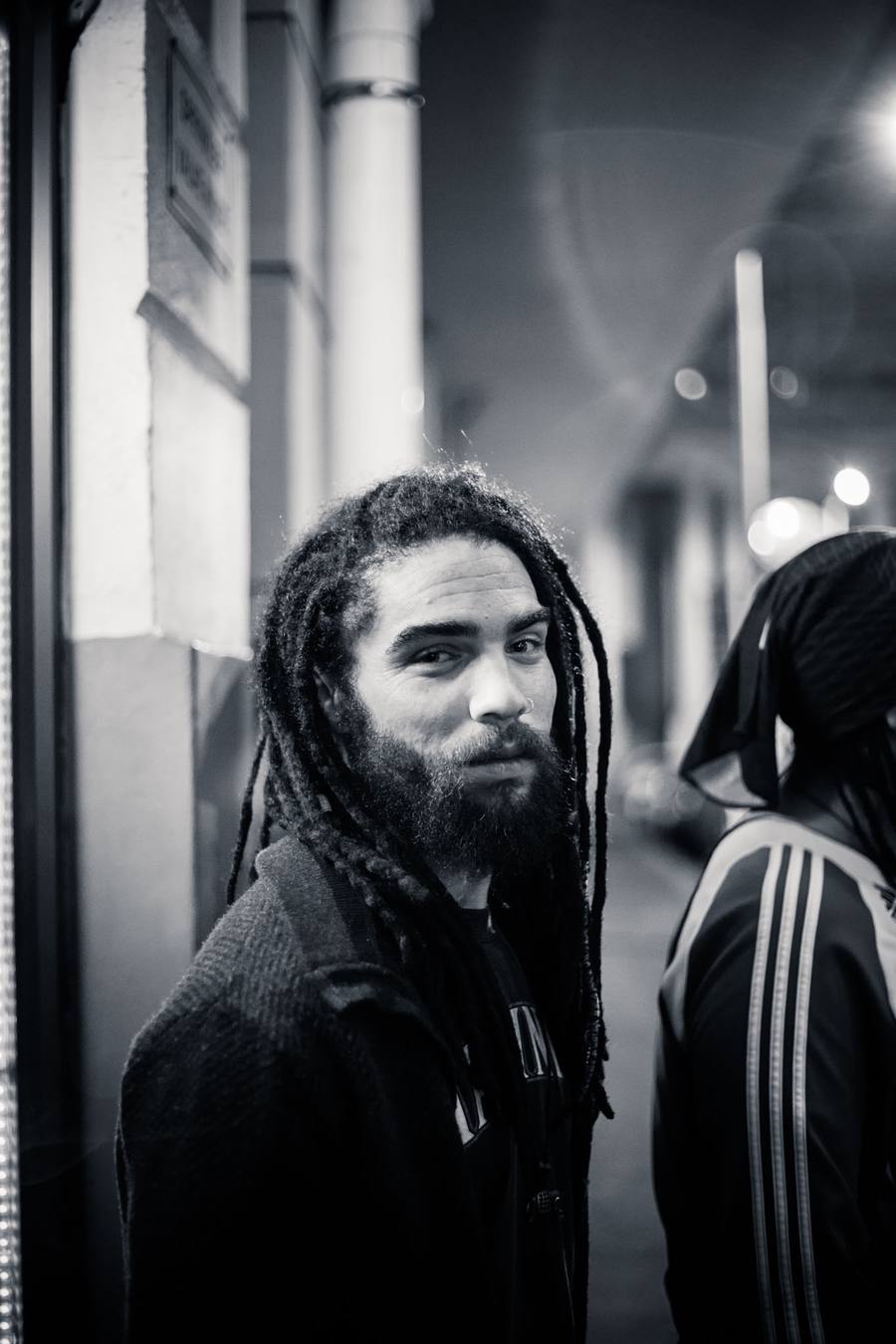 A man with dreadlocks and a beard stands on a street corner at night
