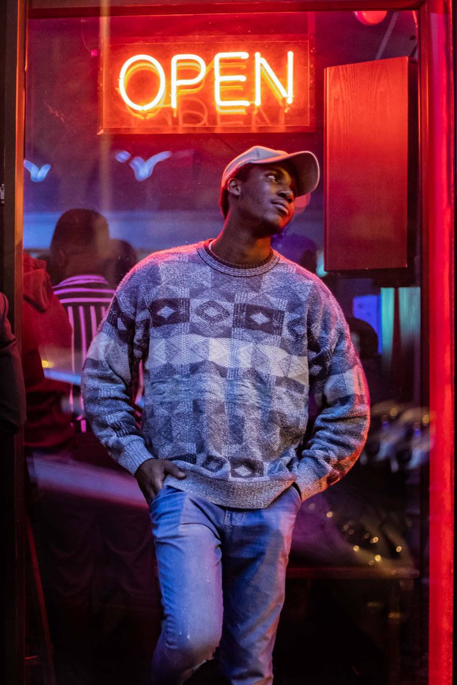Man in patterned jumper and cap standing outside a bar with neon sign