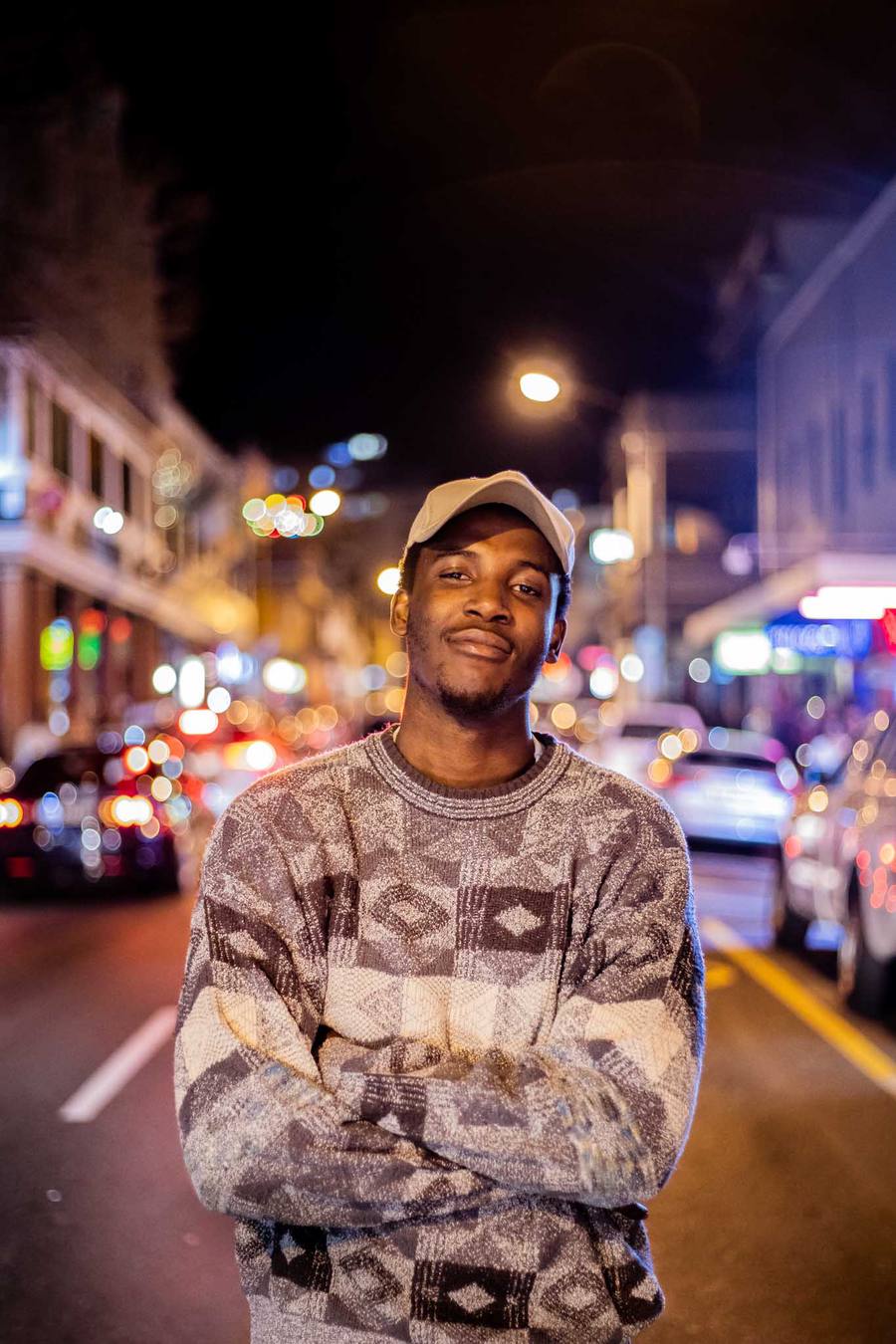 Man in patterned jumper and cap standing on street at night with arms crossed
