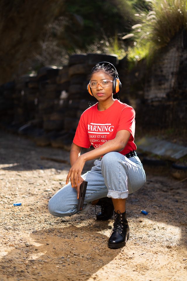 Woman in red shirt and jeans crouching with gun in hand, wearing orange headphones