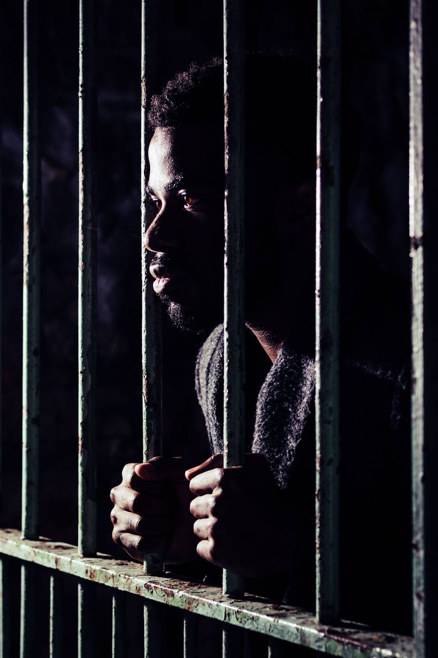 A man with a beard and a jumper holds a metal bar in a dark cell