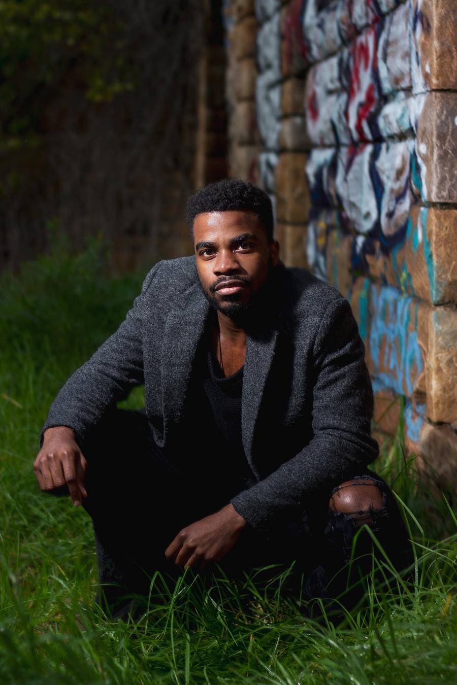 A man in a grey coat sits on grass in front of a graffiti-covered wall