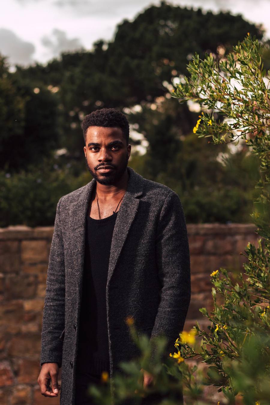 Man in grey coat and black top standing in front of a stone wall and greenery