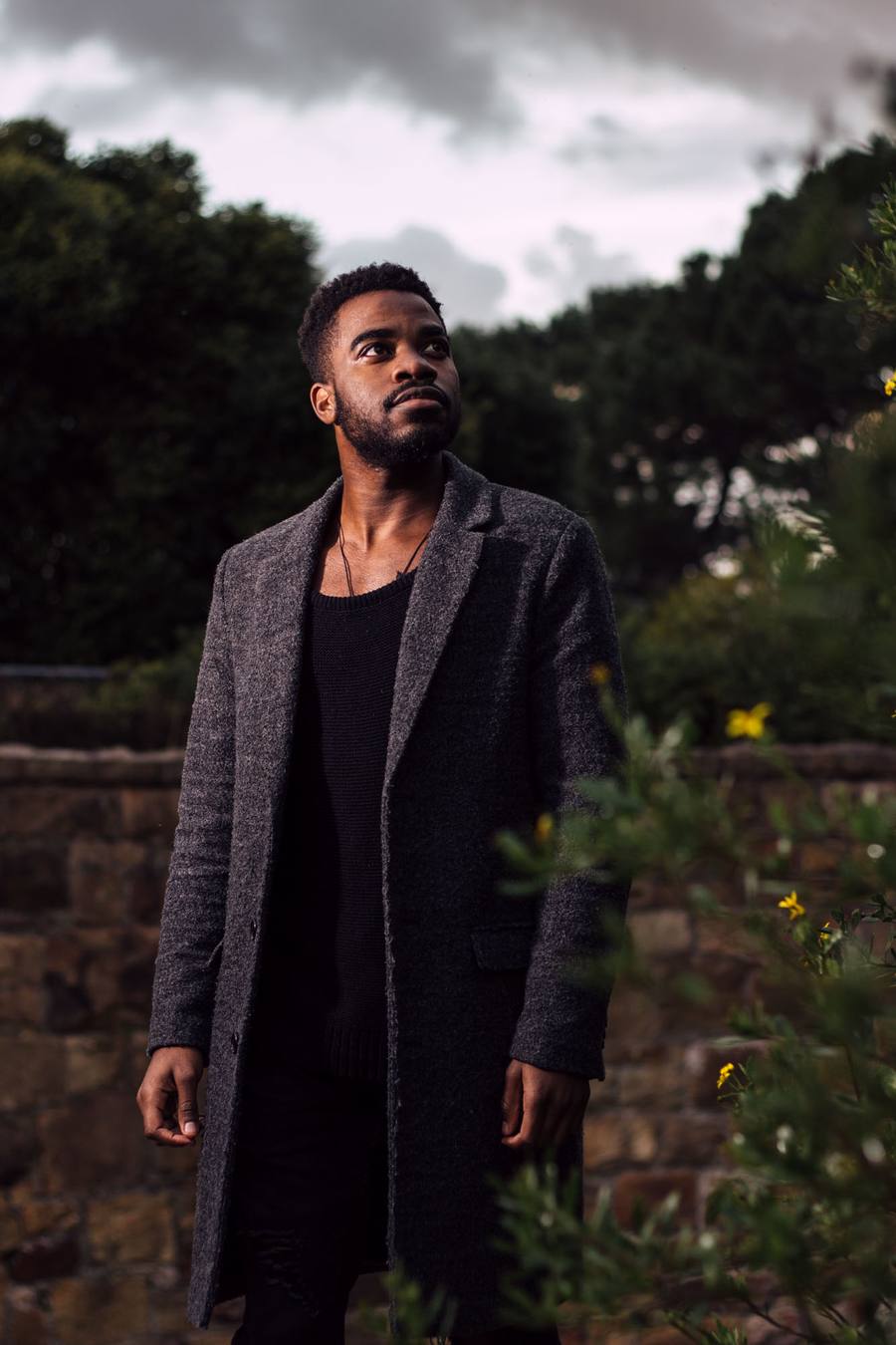 Man in grey coat and black top standing in front of brick wall and greenery