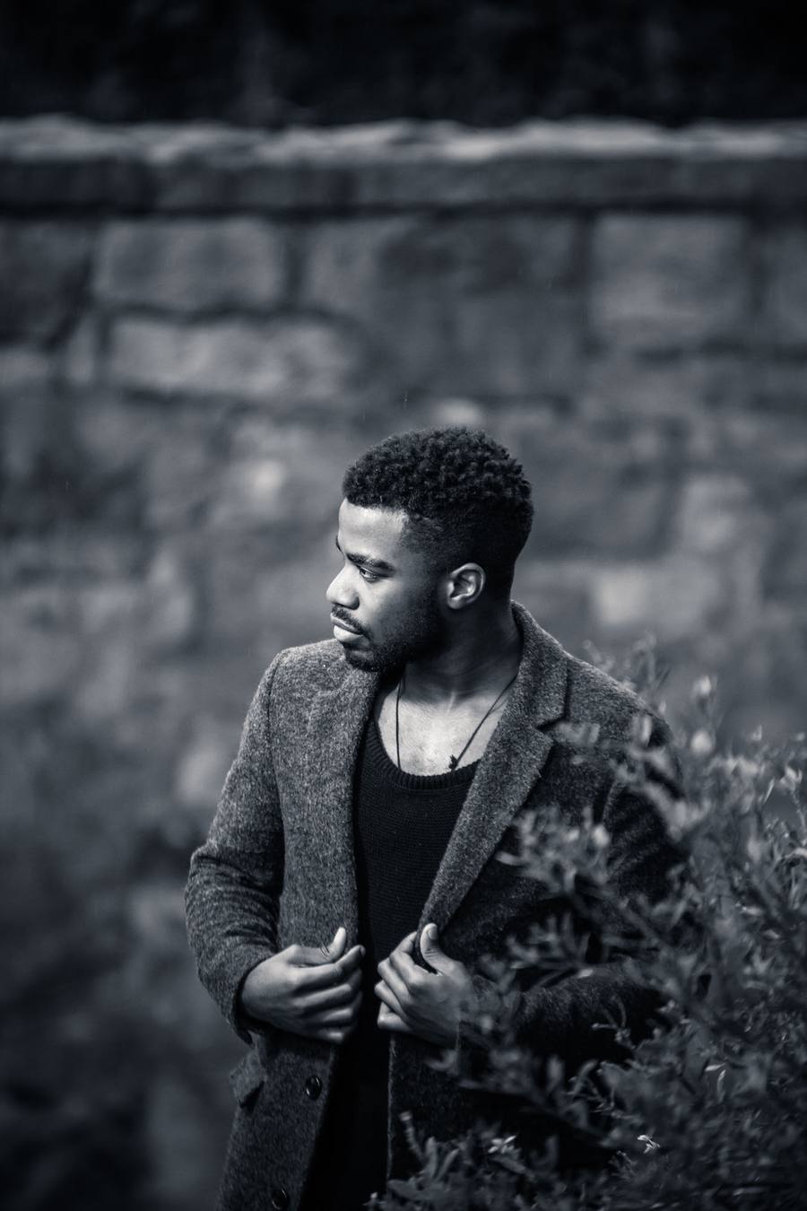 Man in a black shirt and grey coat standing in front of a stone wall