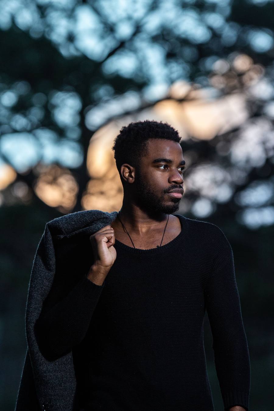 A man in a black top and grey jacket stands in a forest, looking to the side.