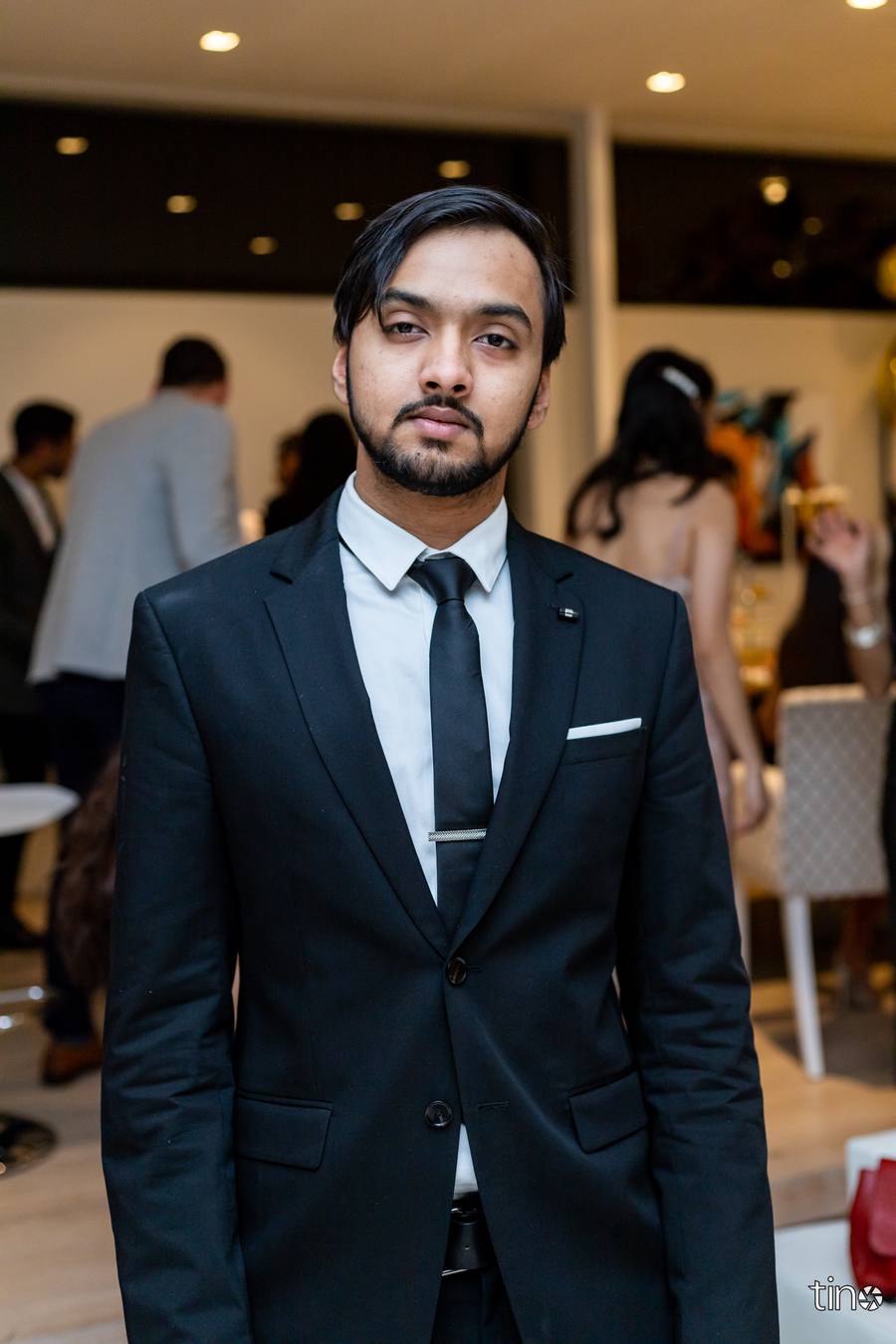 Man in a black suit and tie stands in a room with other people