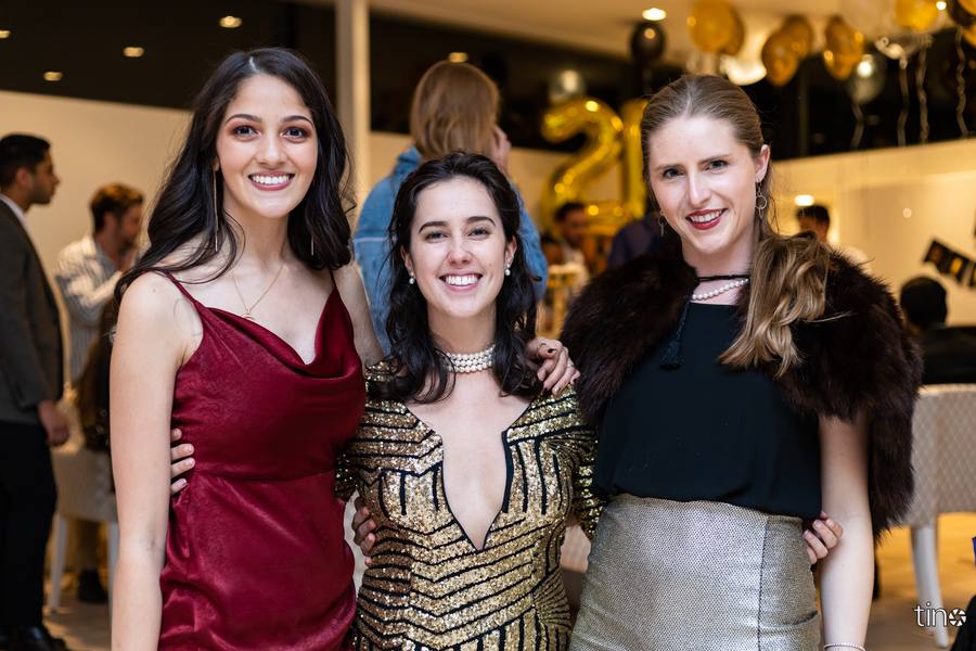 Three women smiling at camera, one wearing red dress, another gold sequins, third in fur jacket