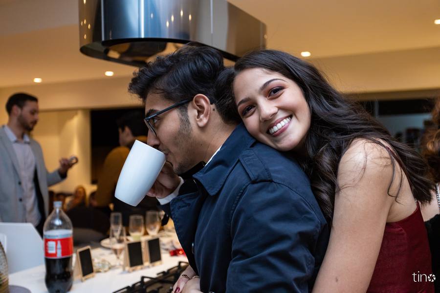 A man drinks from a cup while a woman hugs him from behind in a kitchen.