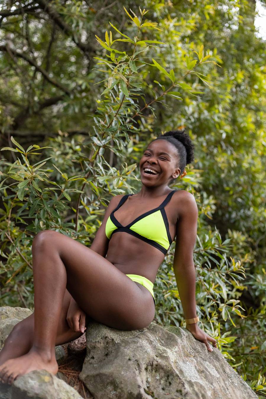 A woman in a yellow and black bikini sits on a rock, smiling, with greenery behind her.