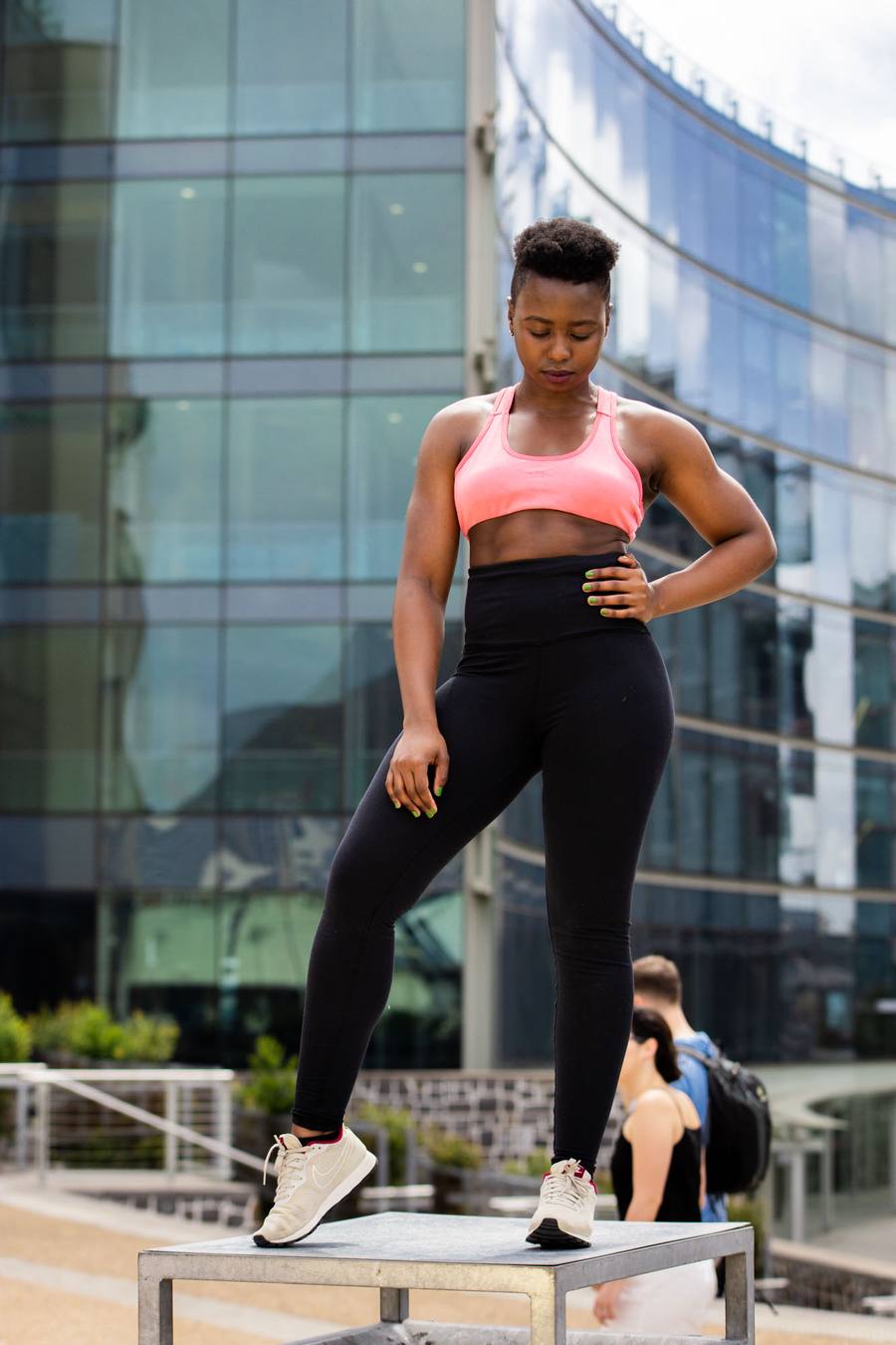 Woman in pink sports bra and black leggings standing on a bench in front of a building