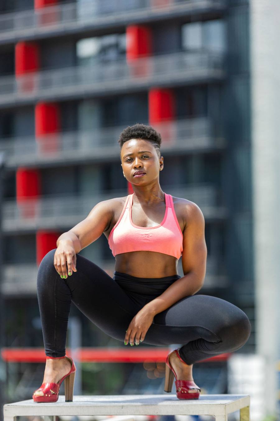 Woman in pink sports bra and black leggings, squatting on a bench in front of a building