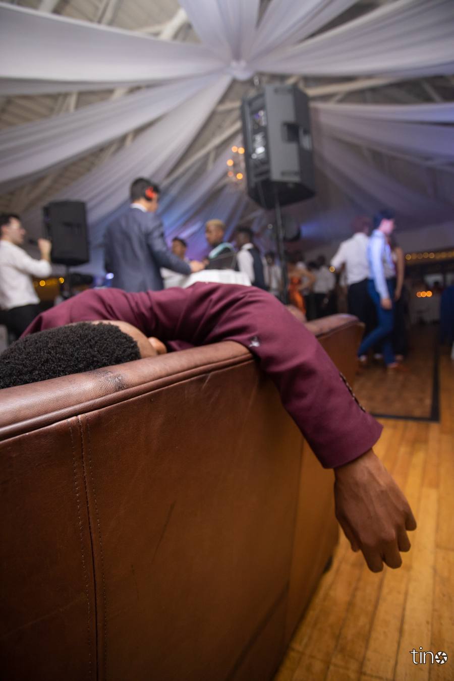 A man sleeps on a sofa while others dance in the background.