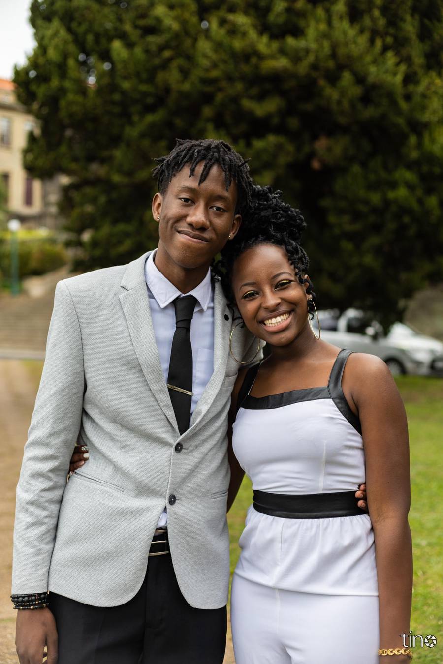 A young couple stands close together, smiling, with a large tree behind them.