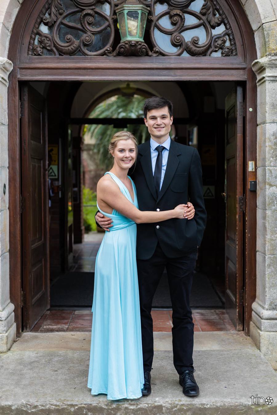 A couple in formal wear stand together, smiling, outside a building.