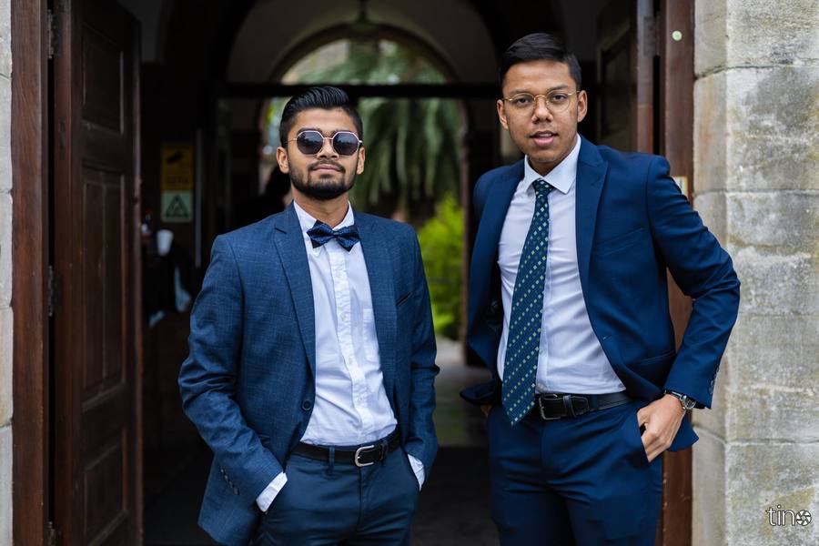 Two men in suits stand outside a building, one with a bow tie and the other with a tie.