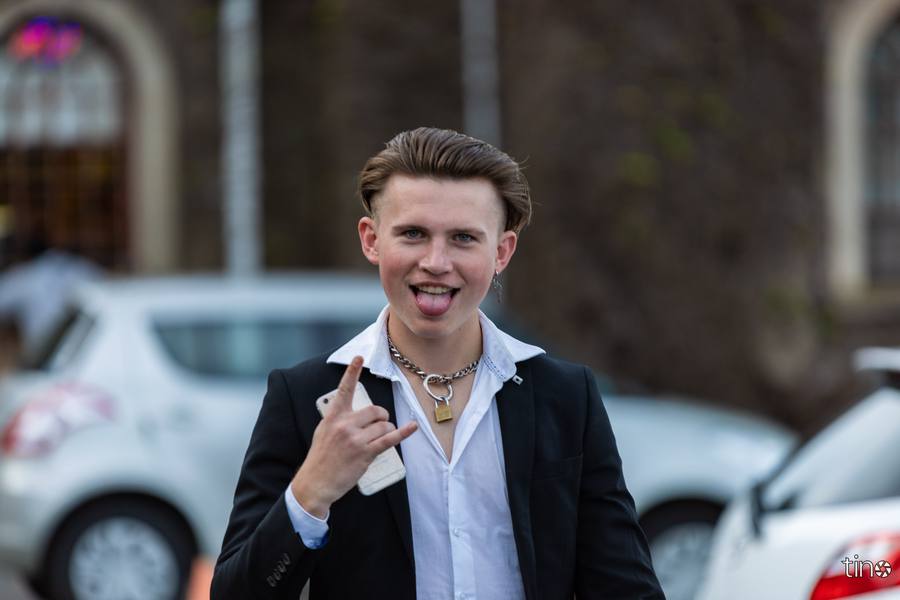 A young man in a suit and chain necklace gives a peace sign with his tongue out