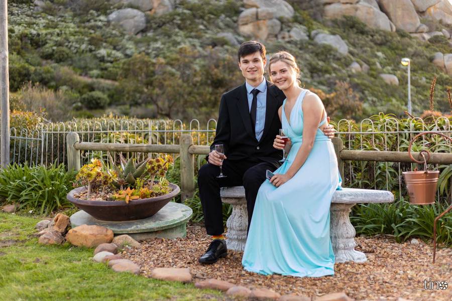Couple in formal attire sitting on a bench in a garden with a rocky backdrop.