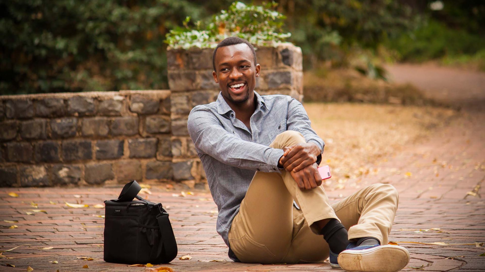 A man sits on a brick path, smiling, with a camera bag beside him.