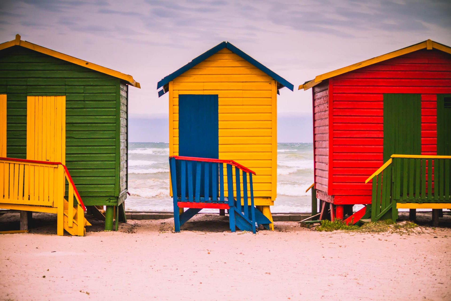 Three brightly coloured beach huts on a sandy beach with the ocean in the background